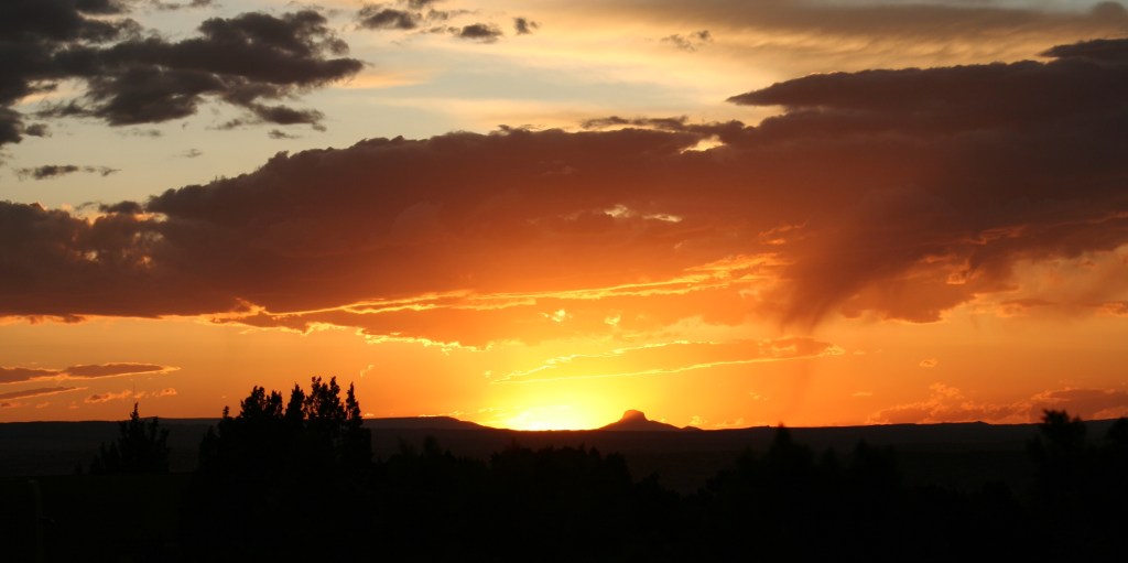 A sunset with the golden sun dipping below the horizon and lighting up dark grey clouds with orange and gold highlights. The bottom of the photo shows a dark horizon with the silhouettes of trees and mountains.